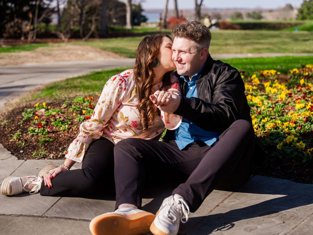 A couple sits together on a curb near a garden of yellow flowers on a sunny day.