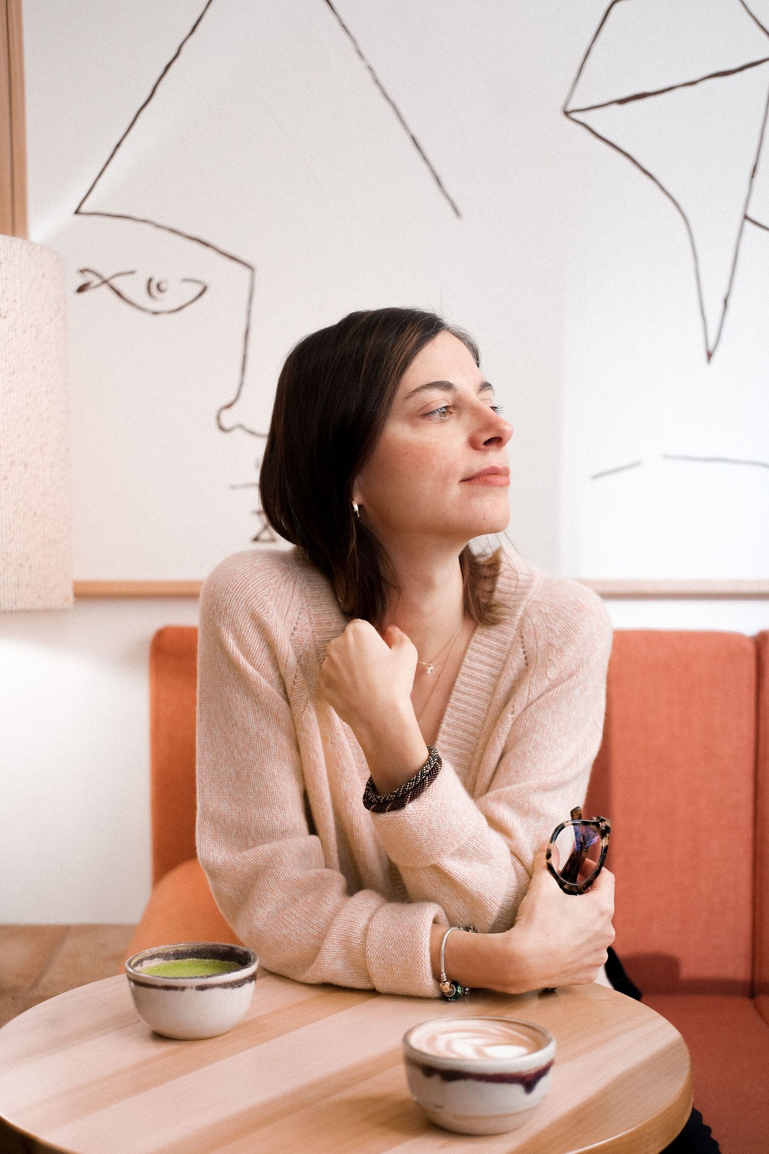 Person in beige sweater sitting at cafe table with matcha and coffee drinks.