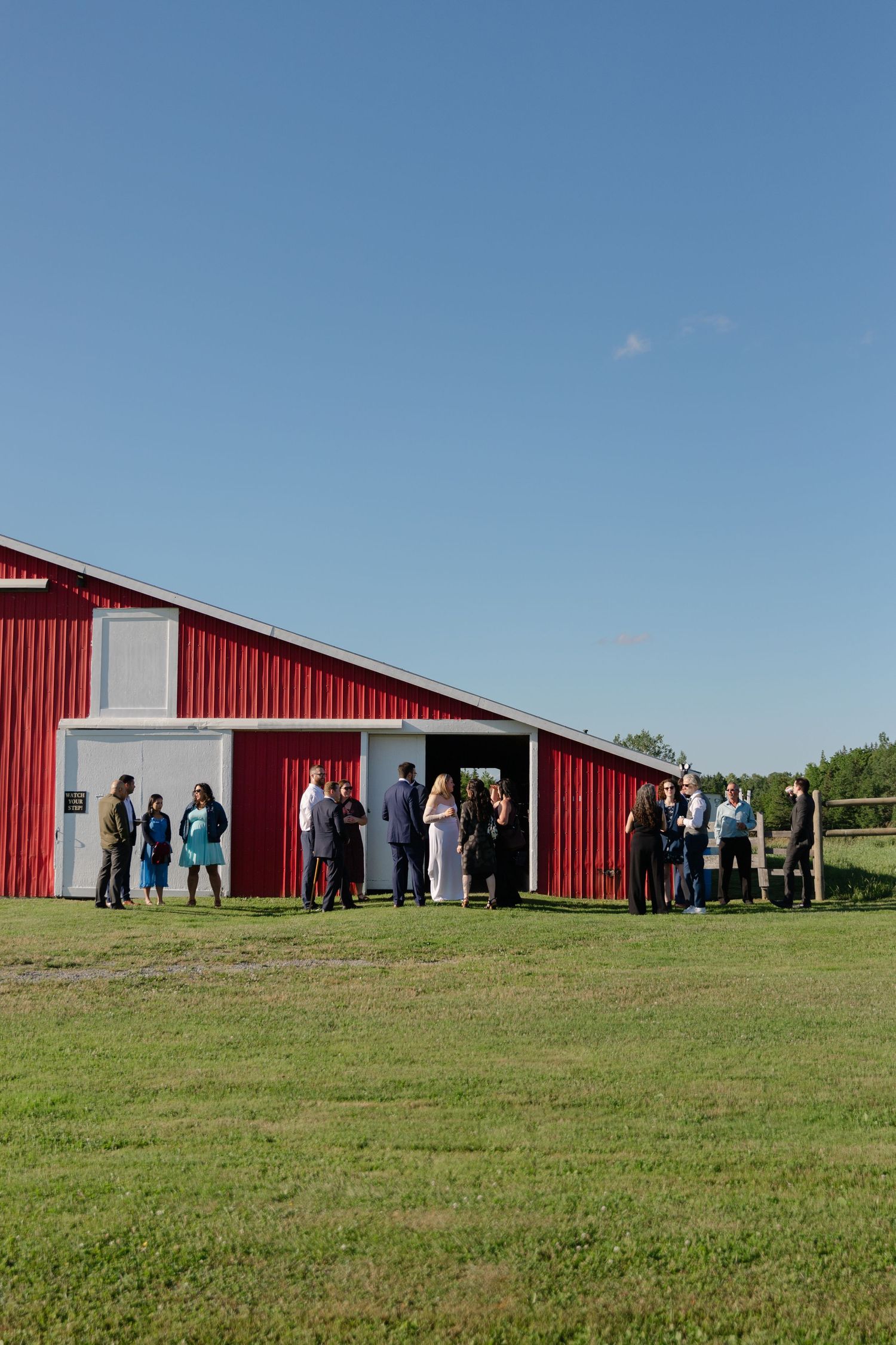 A group gathers outside a red barn on a grassy field under a bright blue sky.