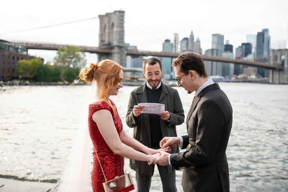 A wedding ceremony takes place by the East River with the Brooklyn Bridge and Manhattan skyline in the background.