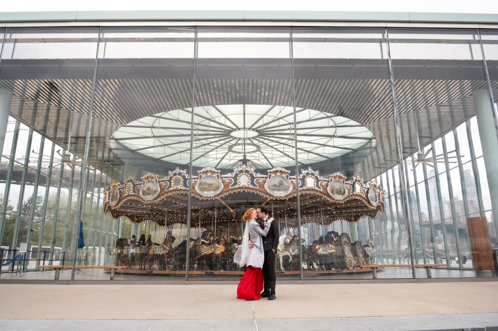 A traditional carousel sits under a modern glass pavilion with two people standing in front of it during daytime.
