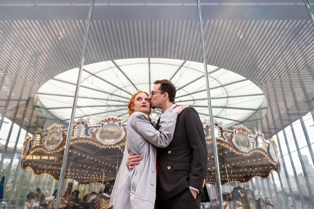 A romantic embrace in front of a vintage carousel under a domed glass ceiling.