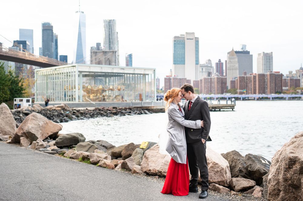 A couple embraces on a rocky waterfront path with the Manhattan skyline and East River in the background.