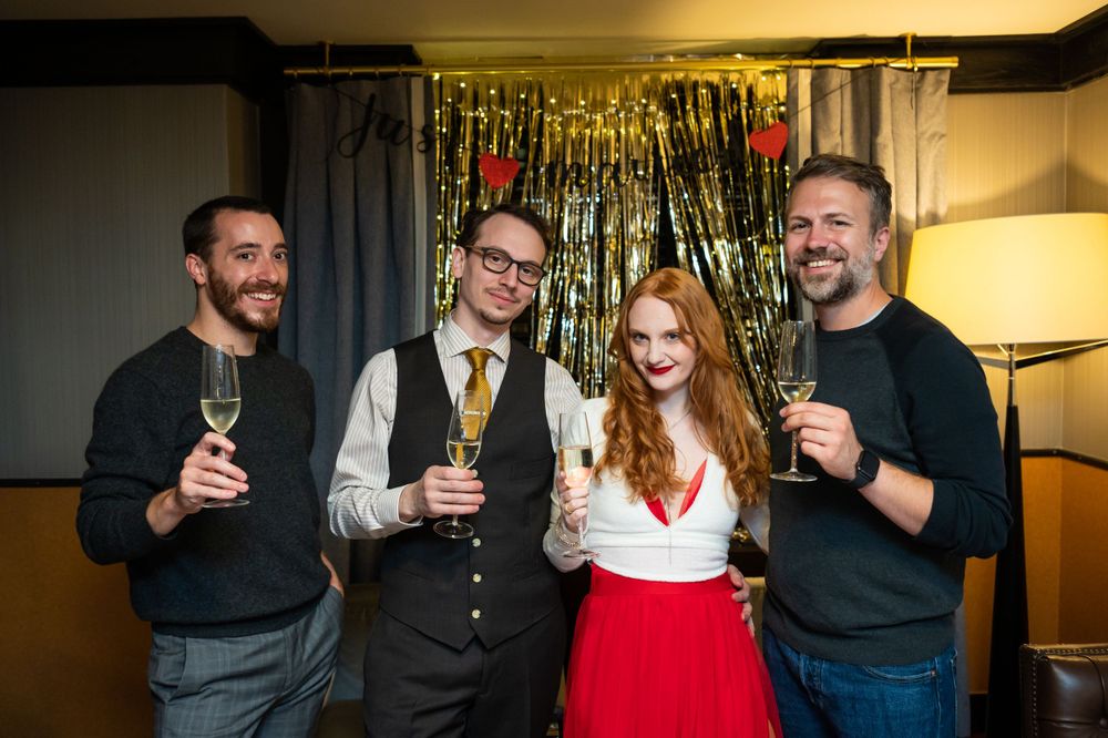 A group of friends celebrating with champagne glasses at a holiday party with sparkly tinsel backdrop.