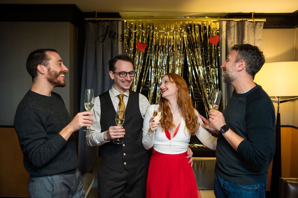 Four friends celebrate at a party with champagne glasses in front of sparkly gold streamers.