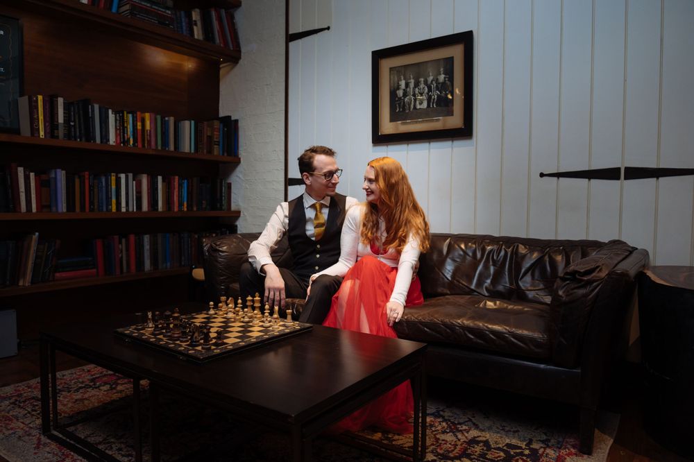 A couple plays chess in a cozy reading room with a leather couch and bookshelves in the background.