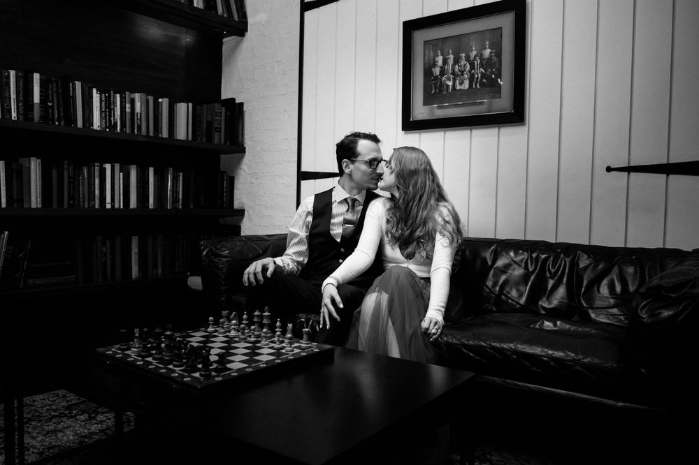 A couple plays chess together in a cozy library room with bookshelves and leather furniture.