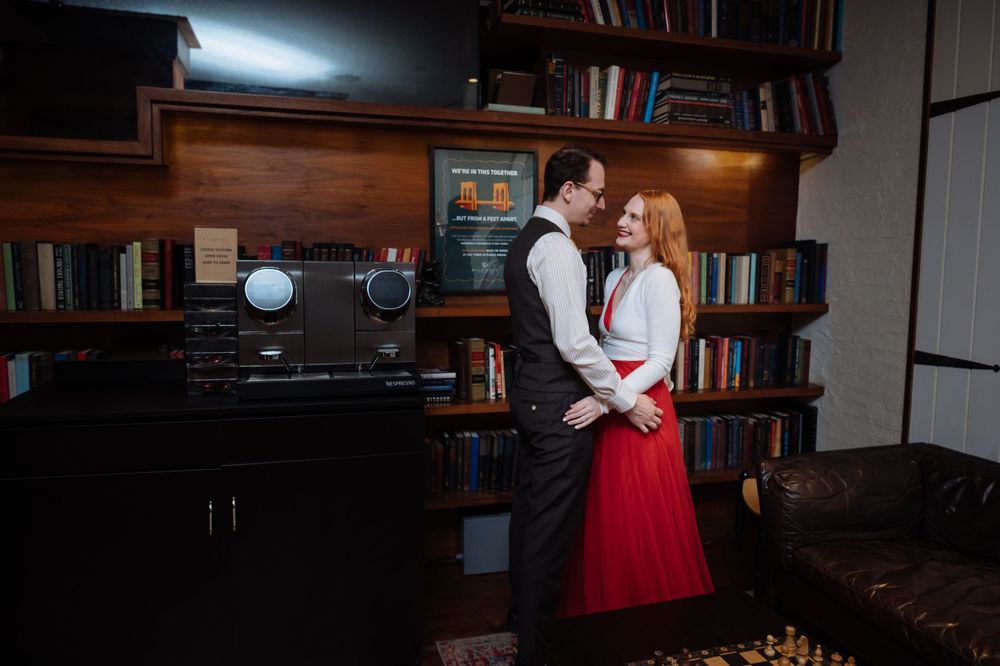 A couple embraces in a cozy library setting with bookshelves and vintage decor, the woman wearing a striking red skirt.