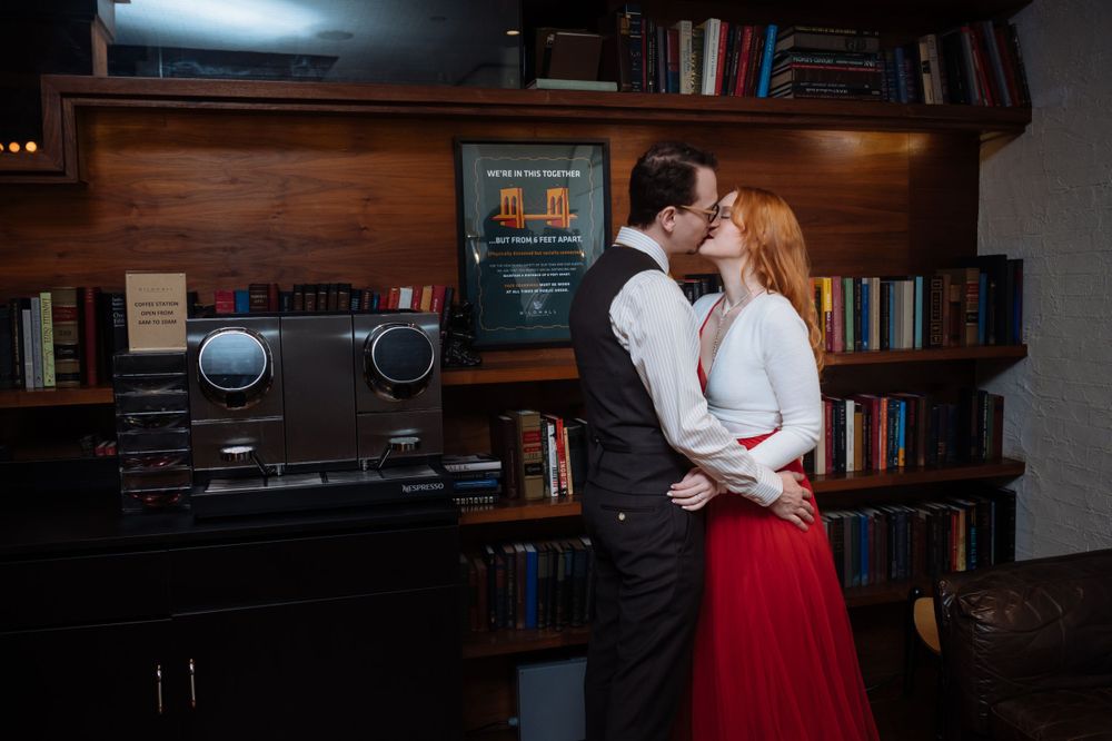 A couple in formal attire embrace in front of a vintage stereo system and bookshelf in a dark, moody setting.