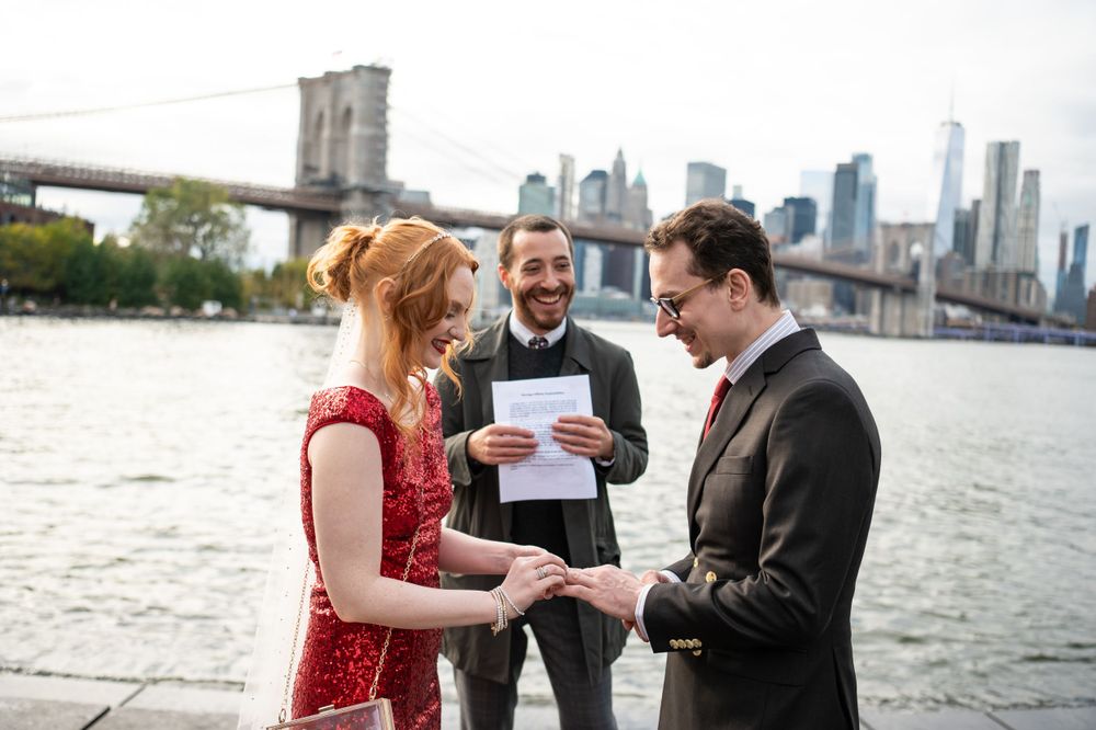 Couple exchanges rings during outdoor ceremony by Brooklyn Bridge and NYC skyline on cloudy day.