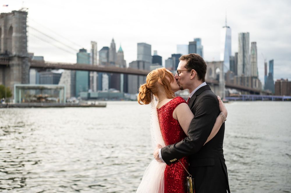 A couple embrace along the East River with the Brooklyn Bridge and Manhattan skyline in the background.