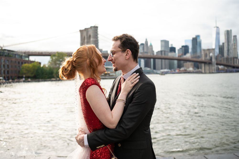 A romantic couple in formal attire embraces by the Brooklyn Bridge and East River in New York City.
