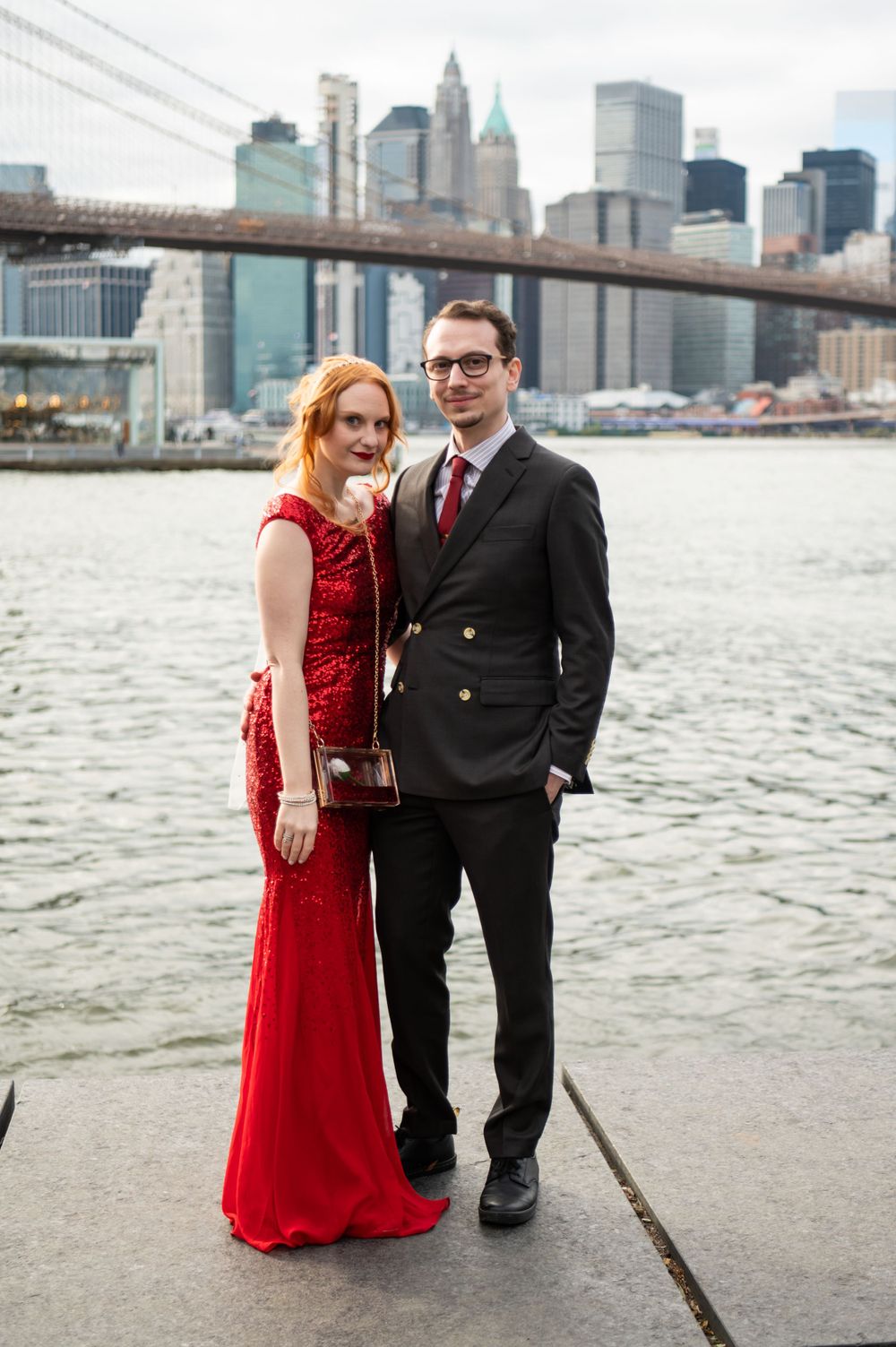Couple in formal wear poses by Brooklyn Bridge with Manhattan skyline in background.