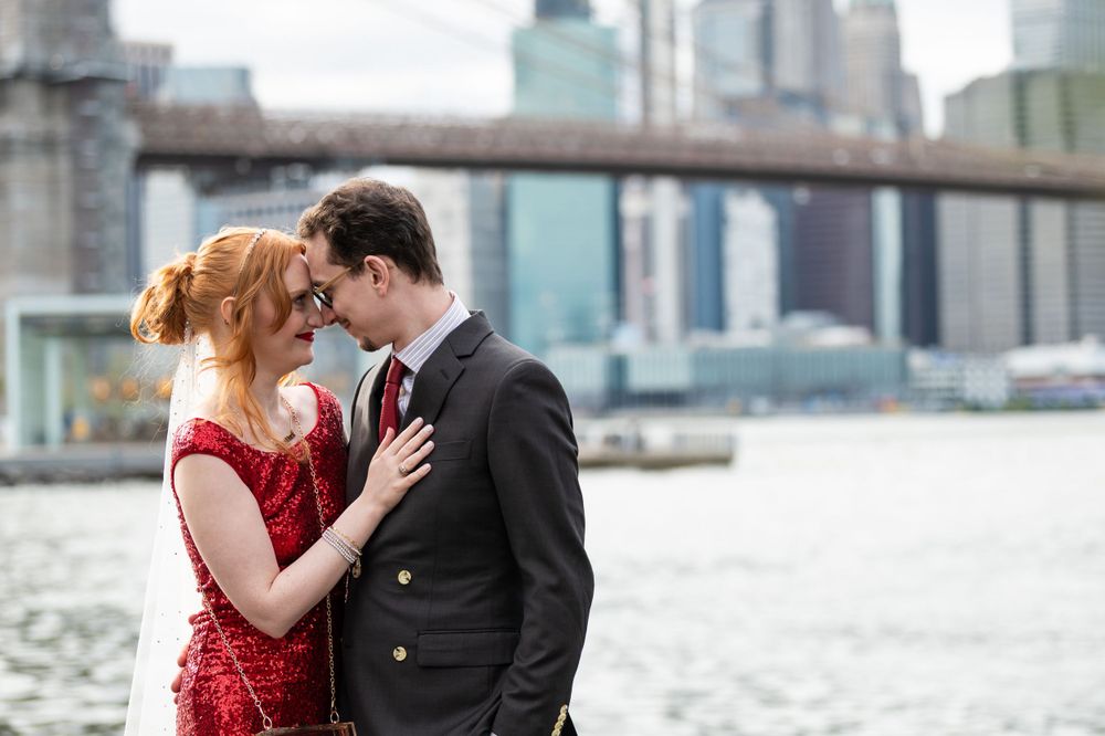 A romantic couple embraces by the waterfront with the Brooklyn Bridge and NYC skyline in the background.