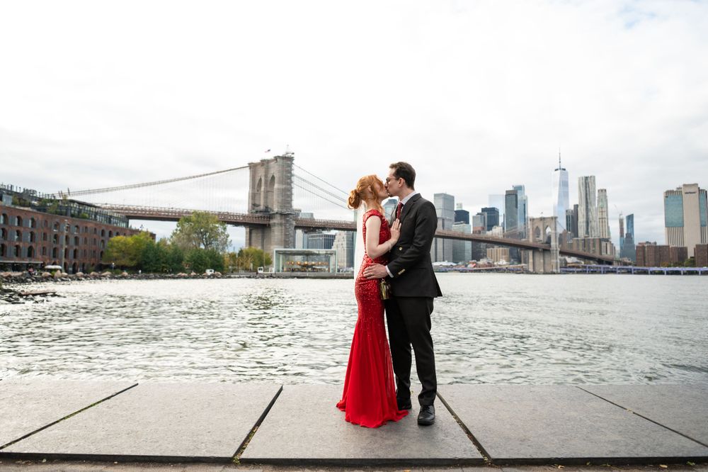 A couple embraces by the Brooklyn Bridge and East River with the Manhattan skyline visible in the background.