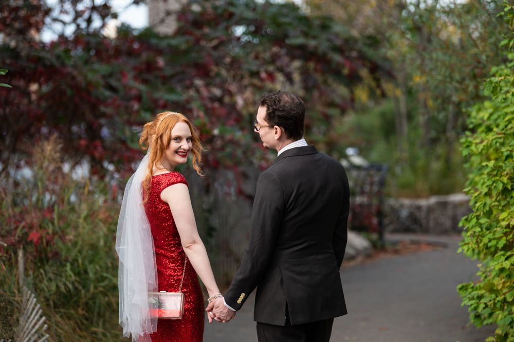 A couple holds hands on a garden path, with the person in a red dress looking back while walking together.