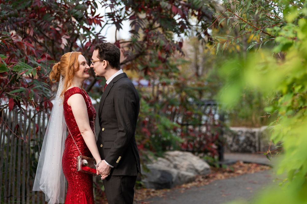 A couple in wedding attire share a tender moment in an autumn garden setting, with red foliage in the background.