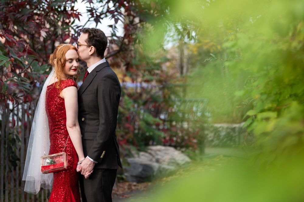 A bride in a sparkly red gown and groom in black suit embrace in a romantic garden setting with green foliage.