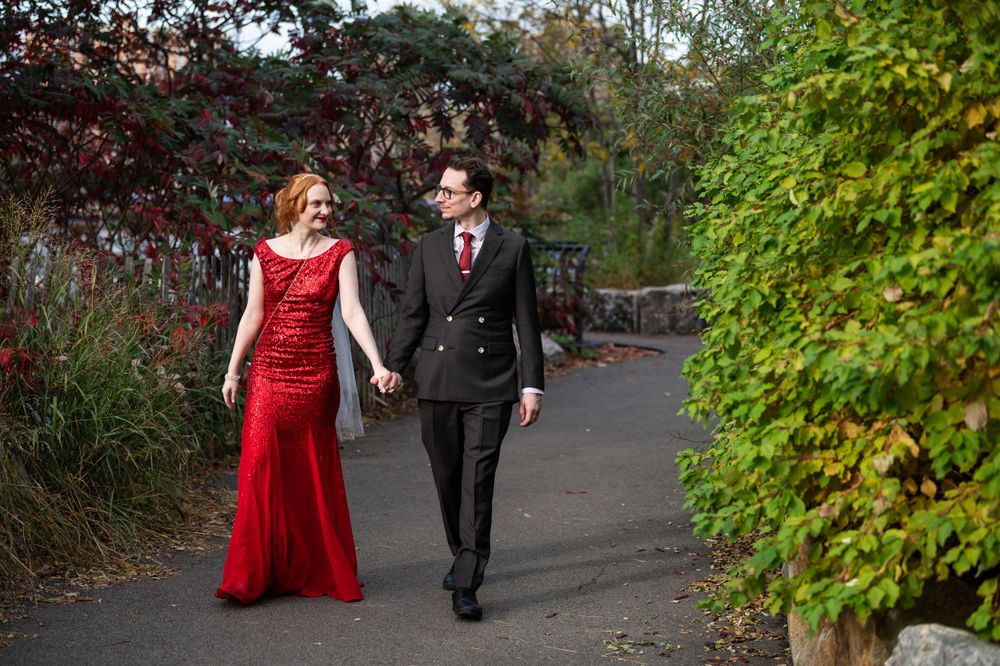 A couple walks hand in hand on a garden path, with the woman wearing a red evening gown and the man in a black suit.