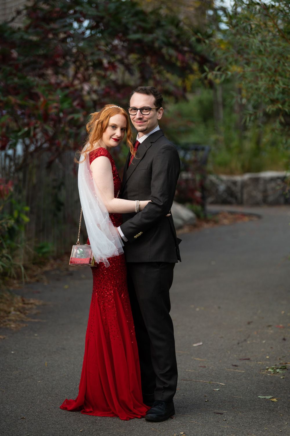 A couple in formal wear embrace on a garden path, with the bride in a stunning red sequined gown and white veil.