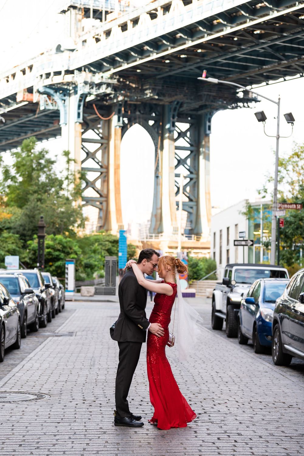 A couple in formal attire embrace on a cobblestone street beneath the Manhattan Bridge in Brooklyn, New York.