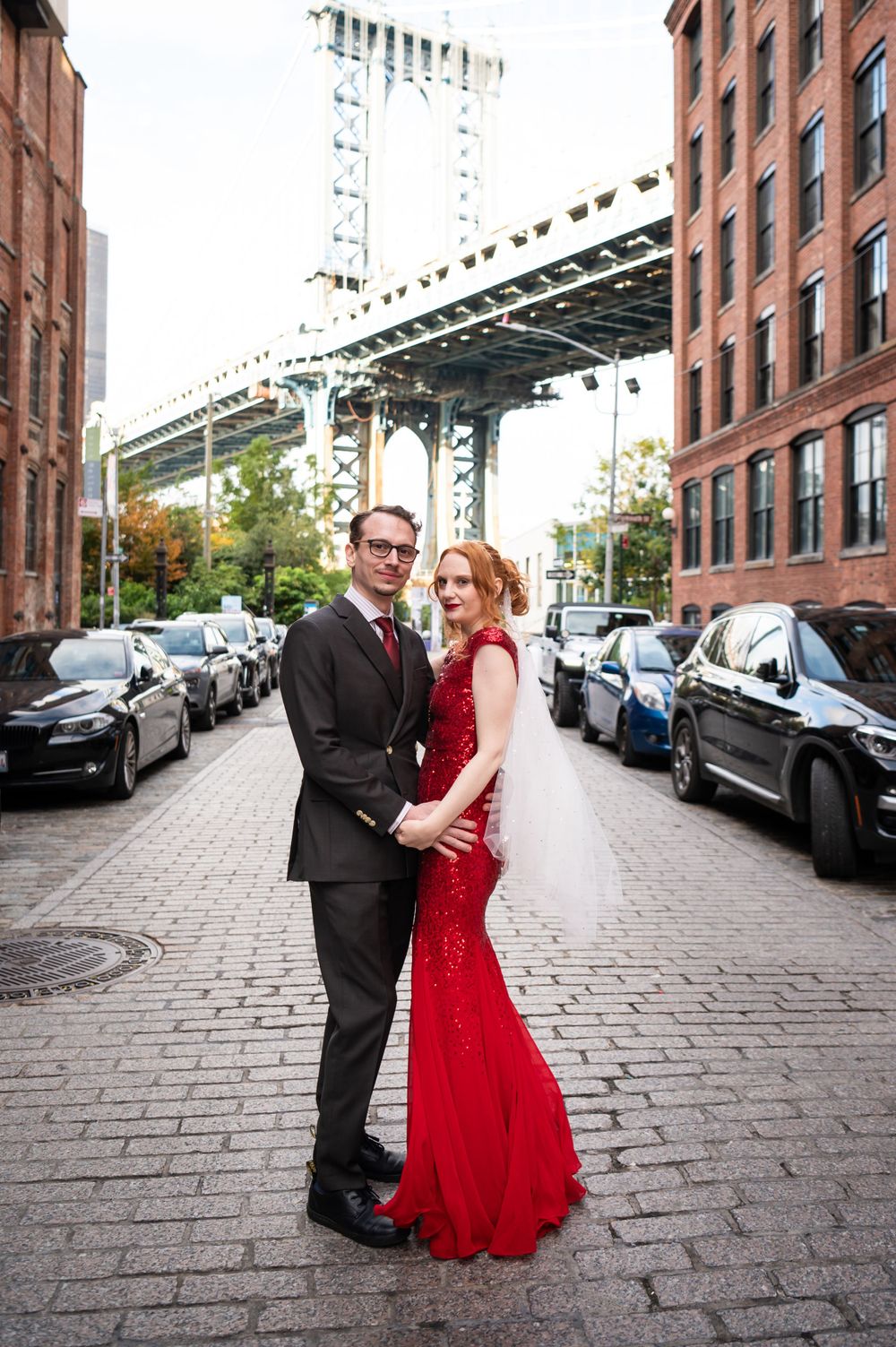 A couple in formal attire poses on a cobblestone street with the Manhattan Bridge in the background in DUMBO, Brooklyn.