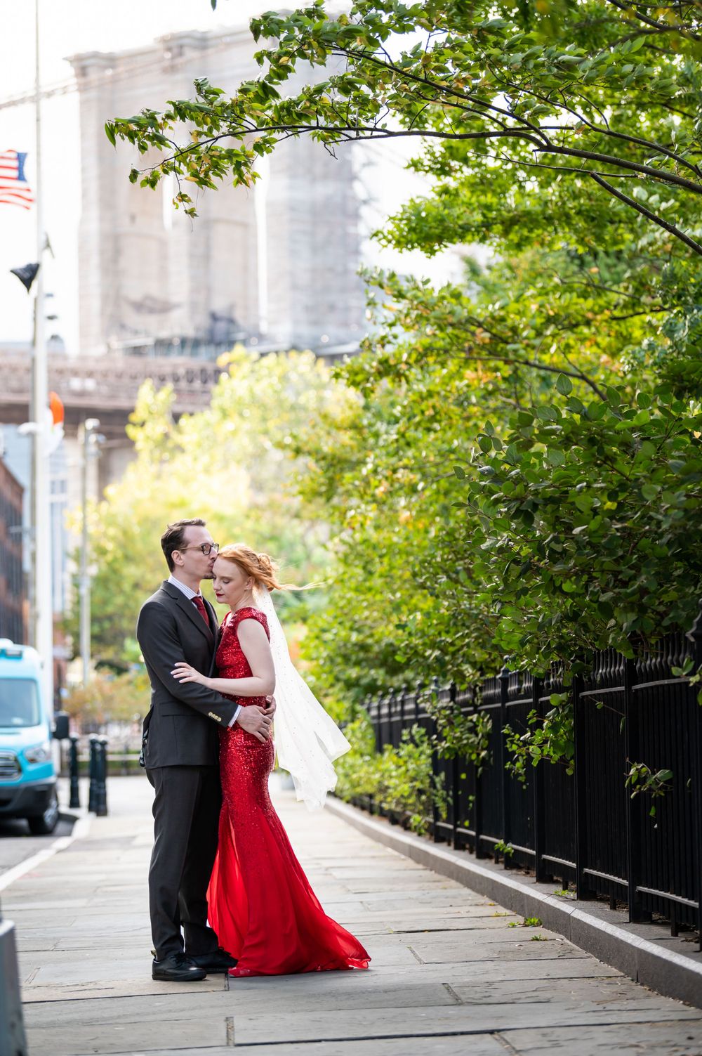 A couple in formal attire embrace on a city sidewalk with lush green trees and urban architecture in the background.
