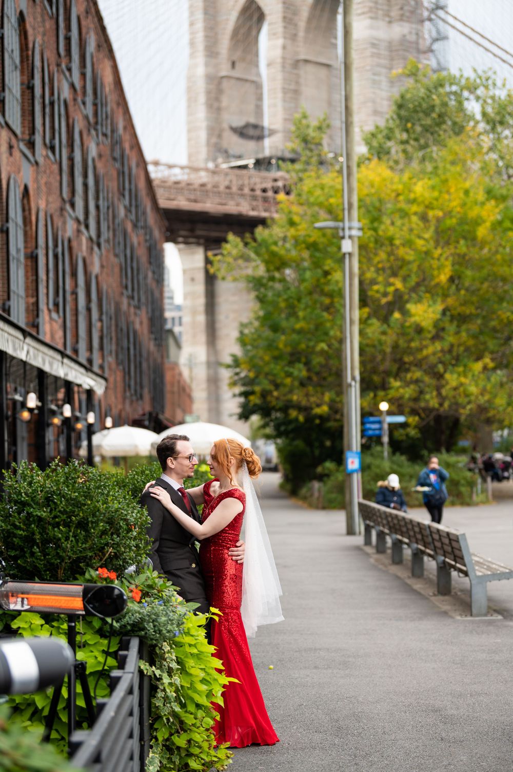 A romantic moment between a couple in formal attire near the Brooklyn Bridge with autumn trees and brick buildings in view.
