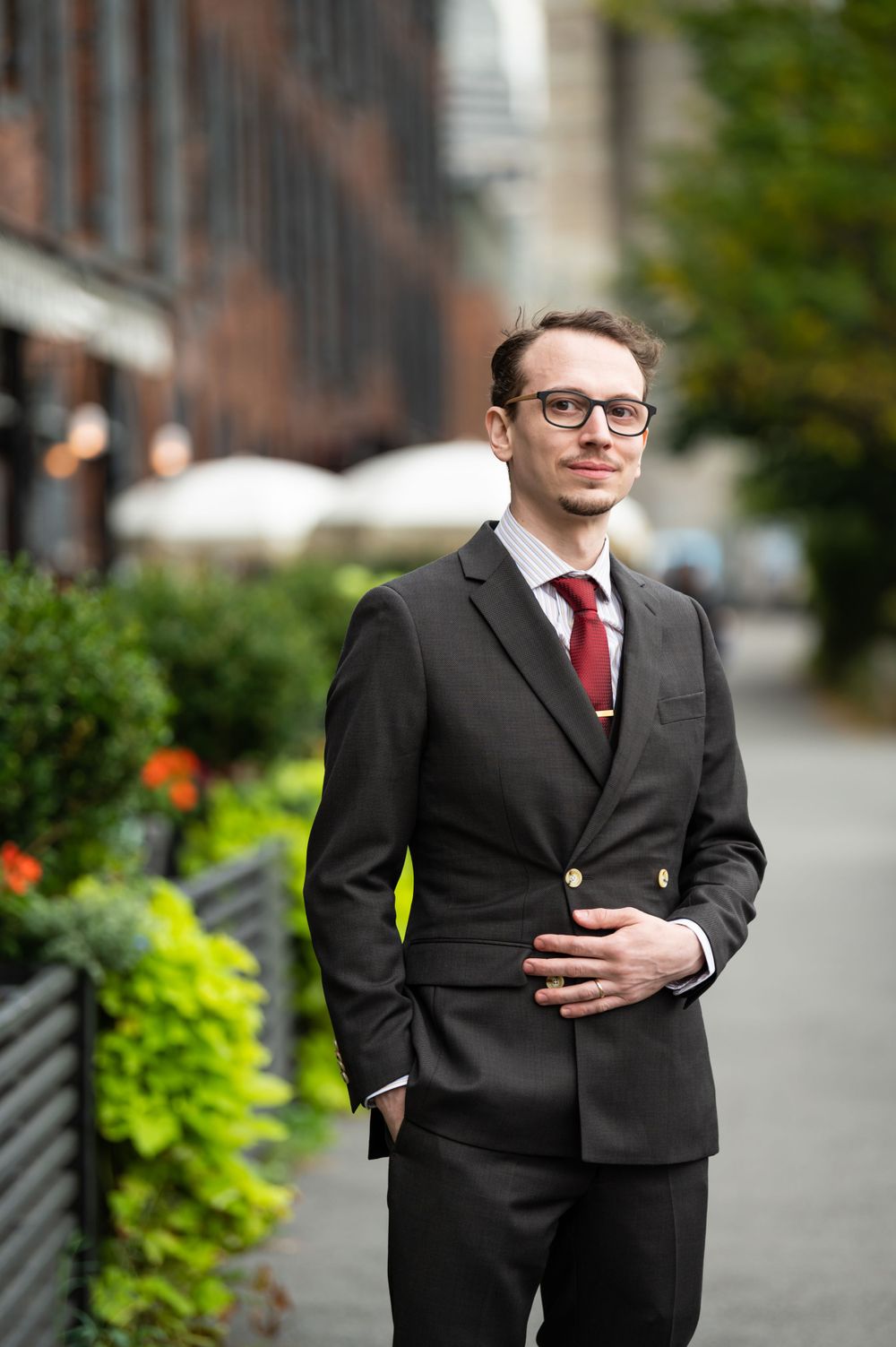A professionally dressed individual in a black suit and red tie stands on a sidewalk near decorative greenery.