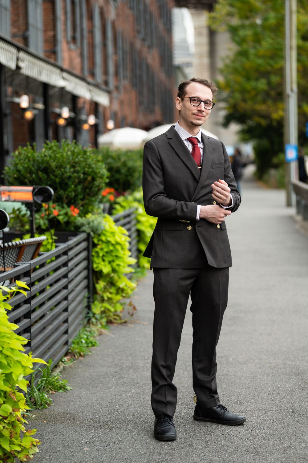 Professional in a dark suit and red tie standing on a city sidewalk next to decorative plants and buildings.