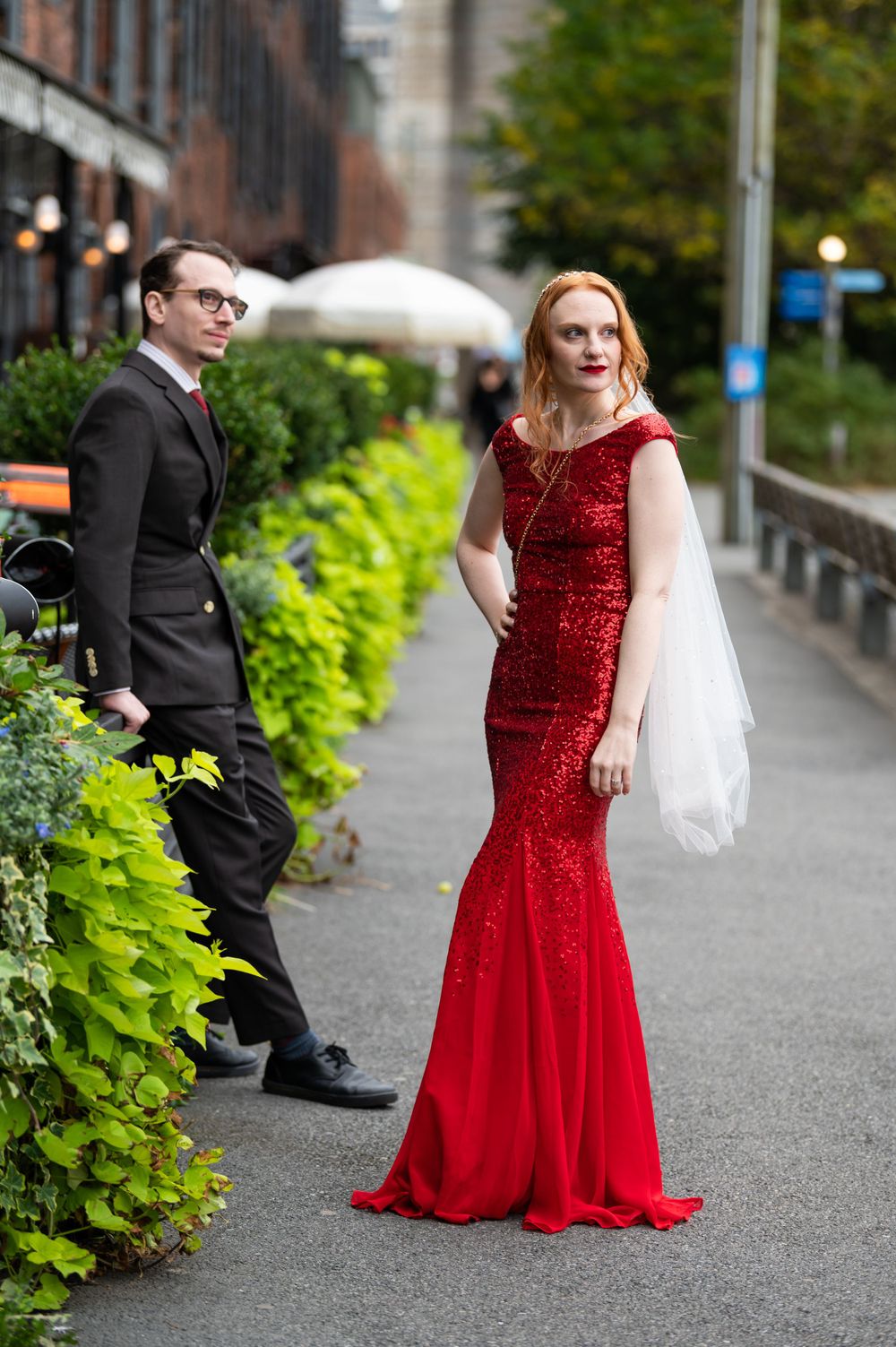 A couple poses on a city sidewalk, with one wearing a black suit and the other in a stunning red sequined evening gown.