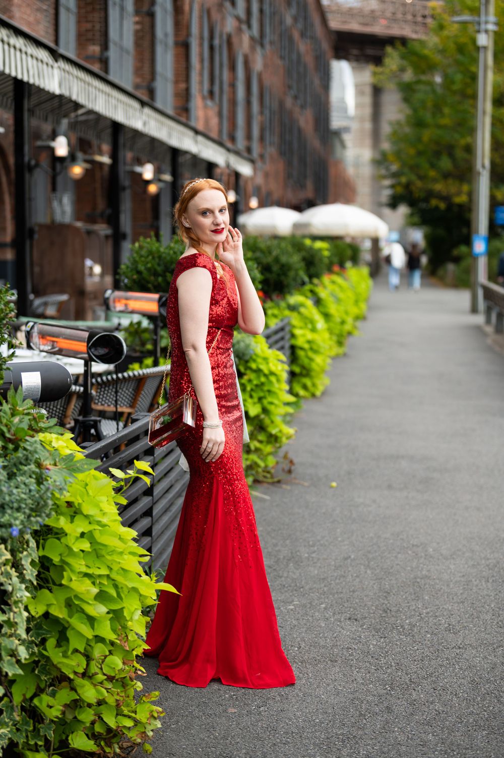 A figure in a stunning red lace gown poses along a walkway with lush greenery and brick buildings in the background.