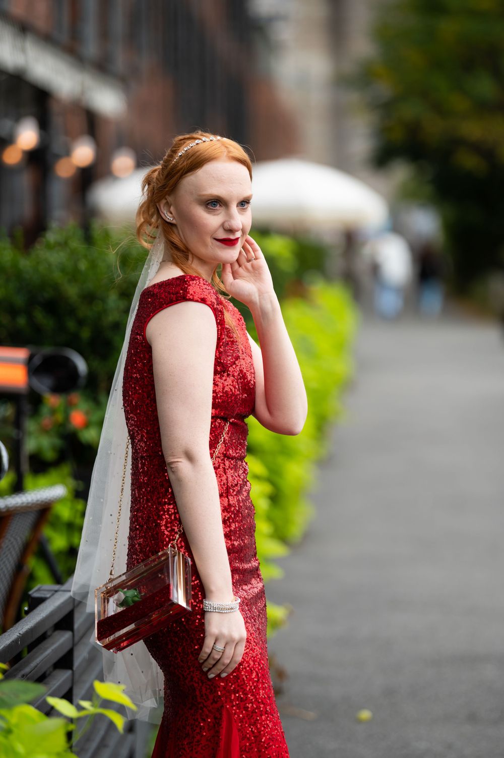 Person wearing a sparkling red evening gown poses outdoors on city street with greenery in background.