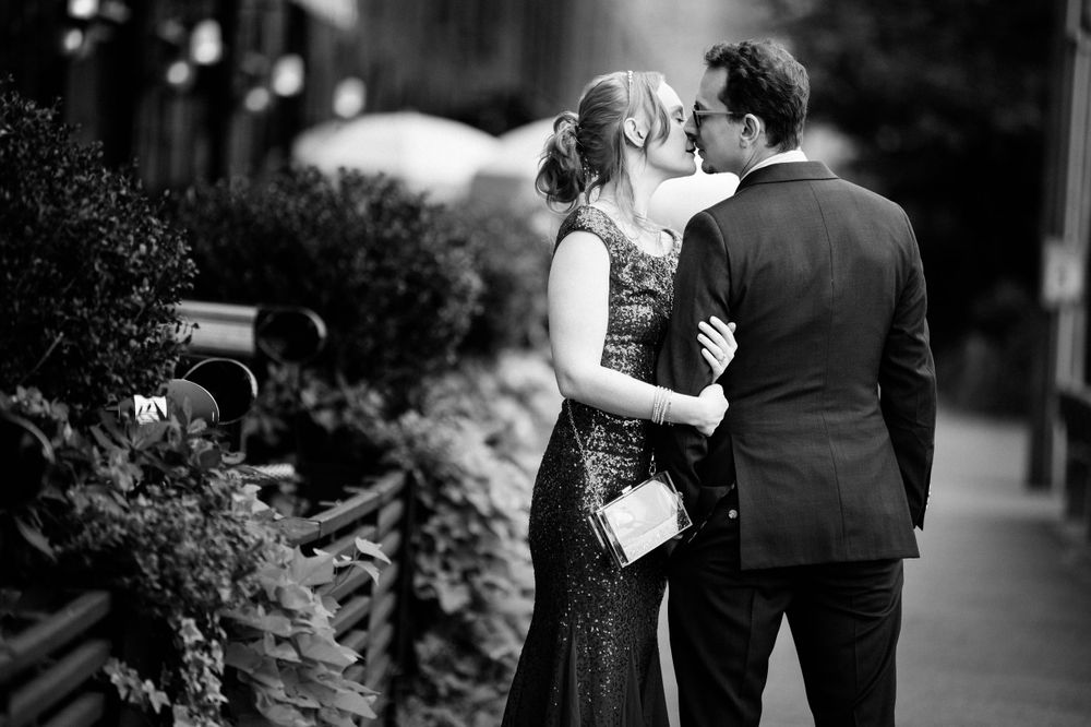 Romantic black and white portrait of a couple sharing an intimate kiss on a sidewalk lined with decorative plants.