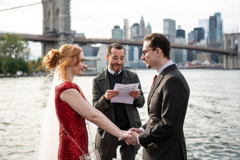 A couple holds hands during their wedding ceremony by the water with the Brooklyn Bridge and NYC skyline in the background.