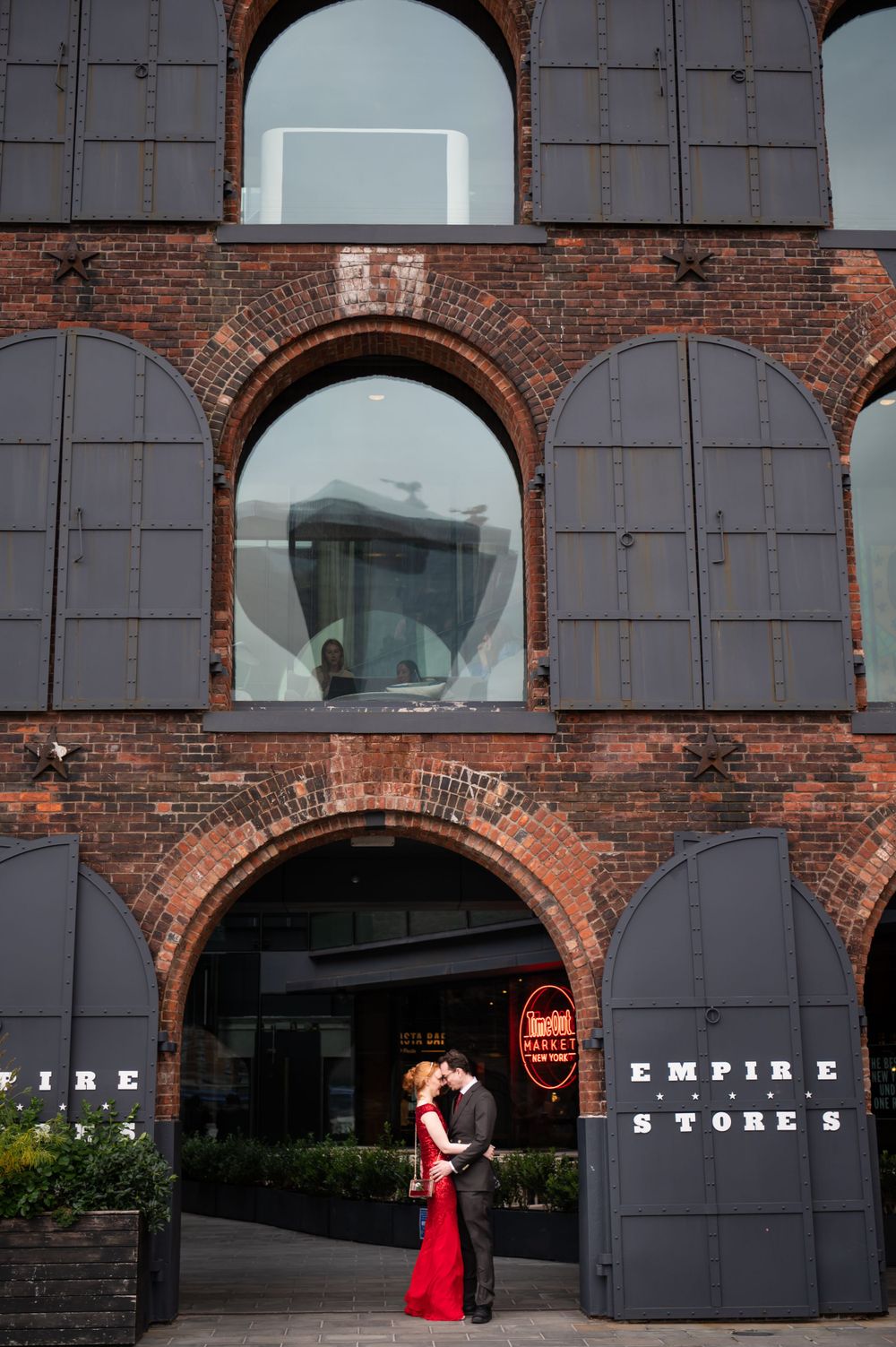 Two people embrace outside the Empire Stores brick building entrance with arched windows and dark metal shutters in DUMBO, Brooklyn.