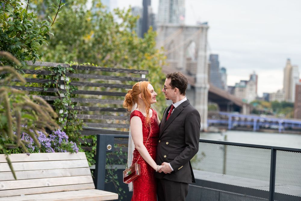 A couple in formal attire stands on a scenic waterfront deck with the Brooklyn Bridge visible in the background.
