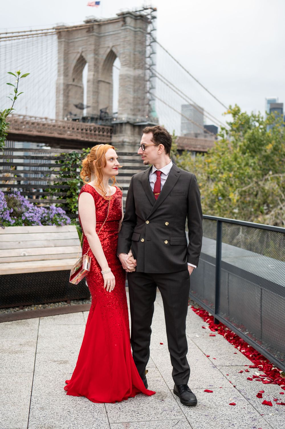 A couple in formal attire poses near the Brooklyn Bridge, with the woman wearing a red gown and man in a dark suit.