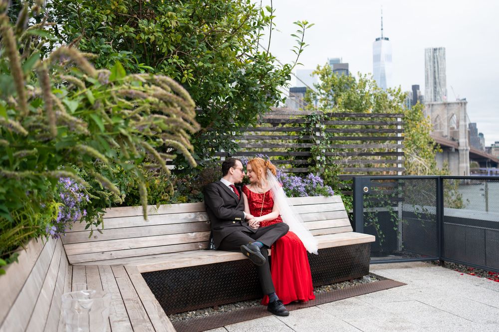 A couple sits on a wooden bench on a rooftop garden with the NYC skyline visible in the background.