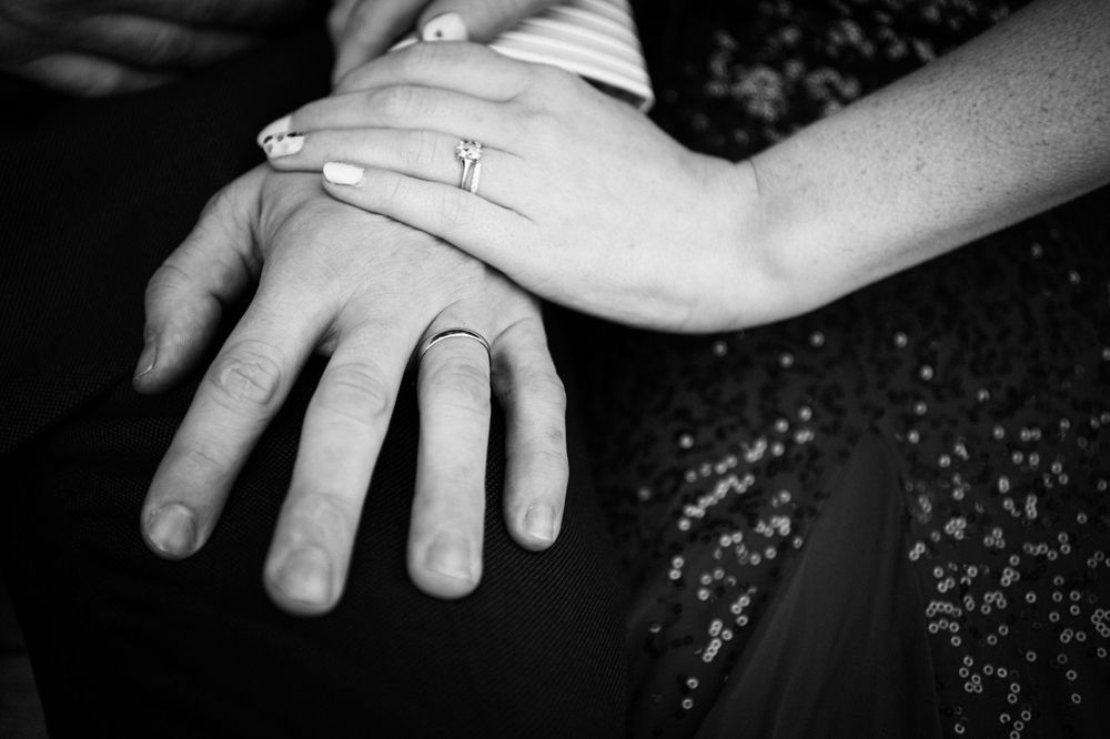 Black and white close-up of hands with wedding bands resting together on a textured surface.