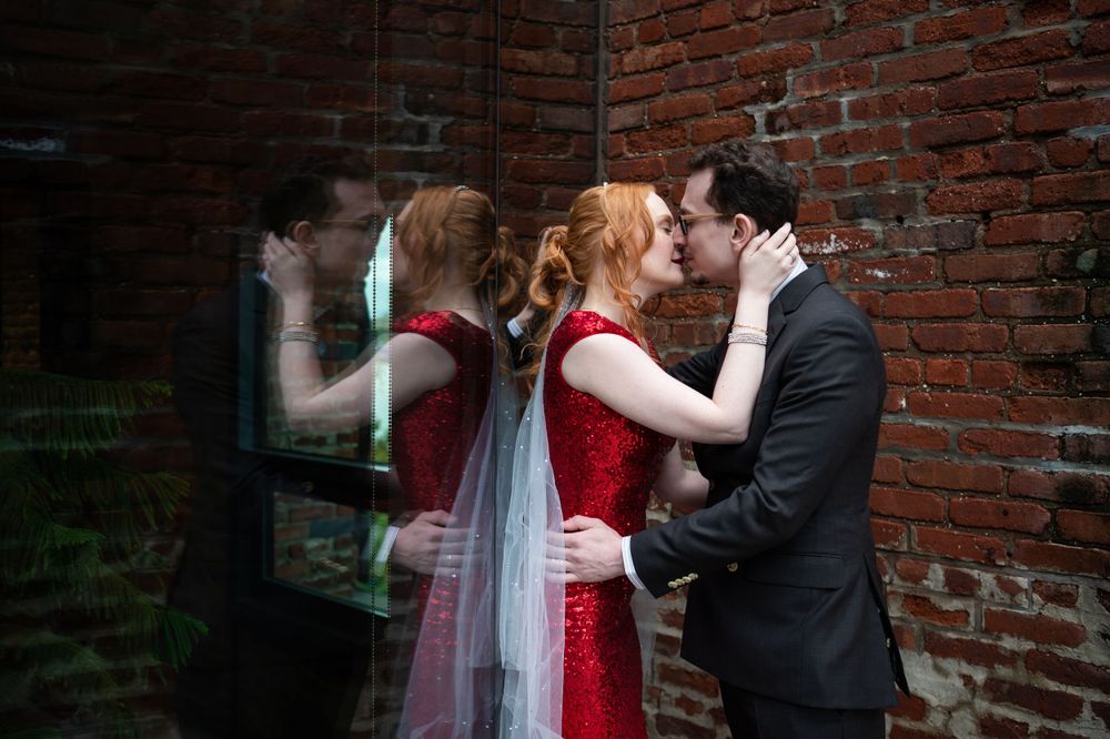 A couple embraces near a brick wall, with the bride wearing a red dress and white veil creating an elegant reflection.