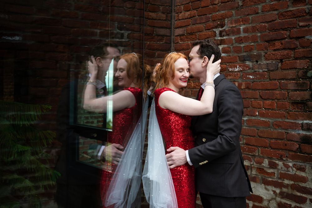 Couple embraces by brick wall, bride wearing sparkling red sequin dress and veil while groom wears classic black suit.