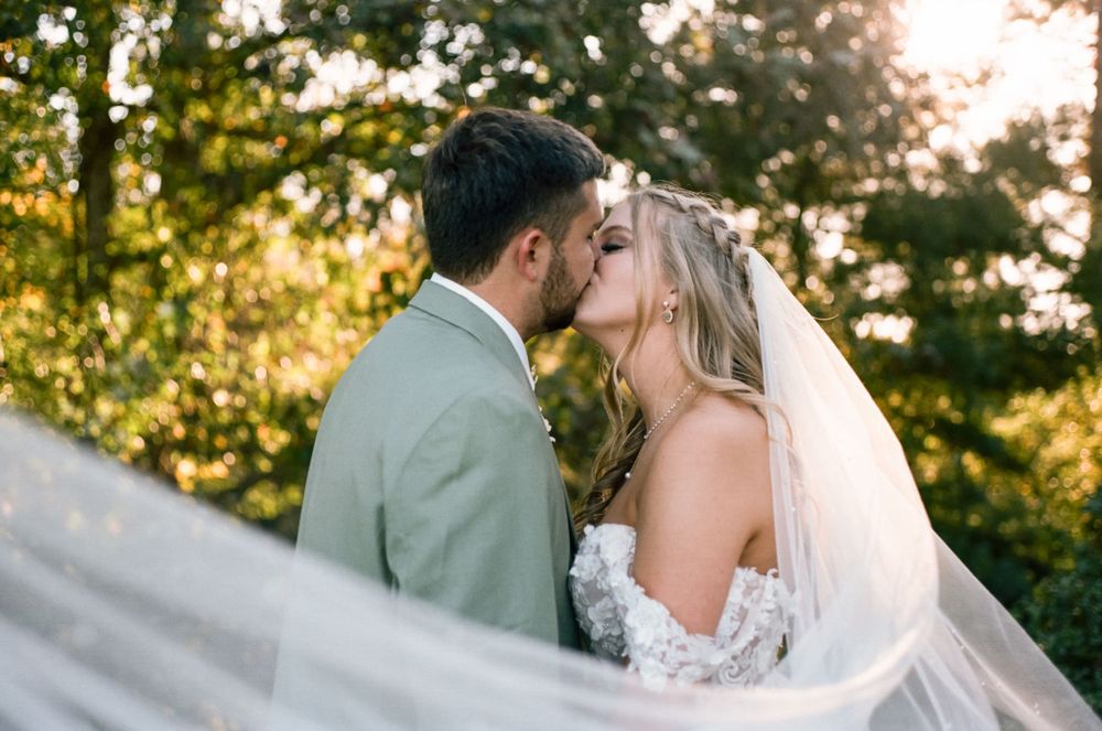 Bride and groom share romantic kiss with flowing veil in golden hour light.