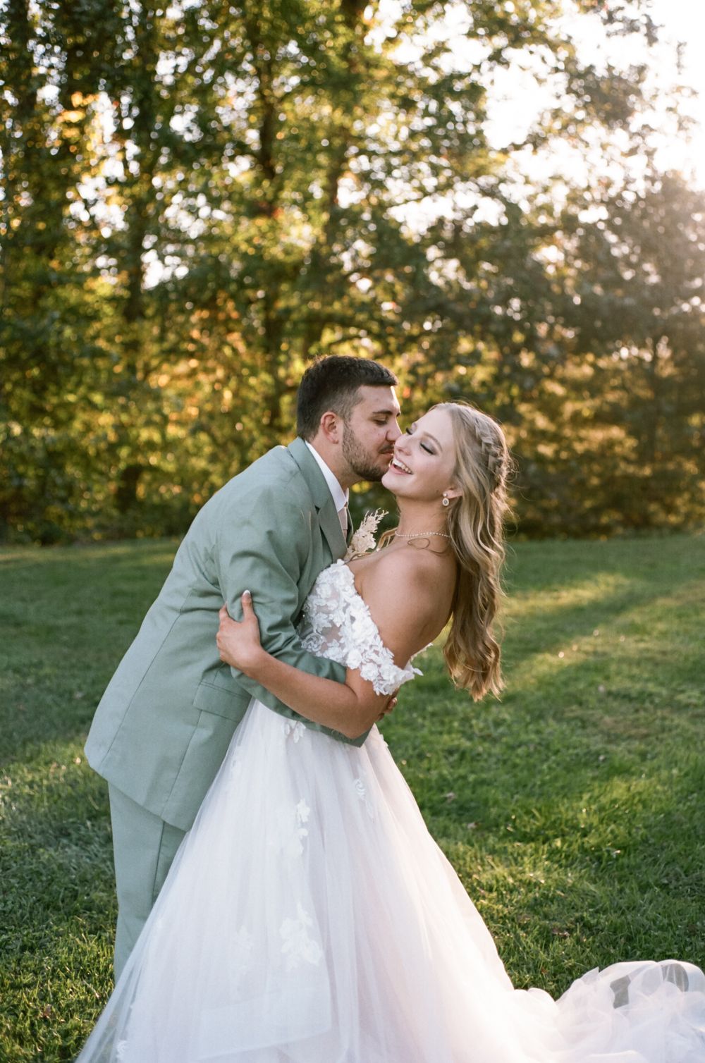 Couple in sage green suit and white wedding dress embrace during outdoor photo session.
