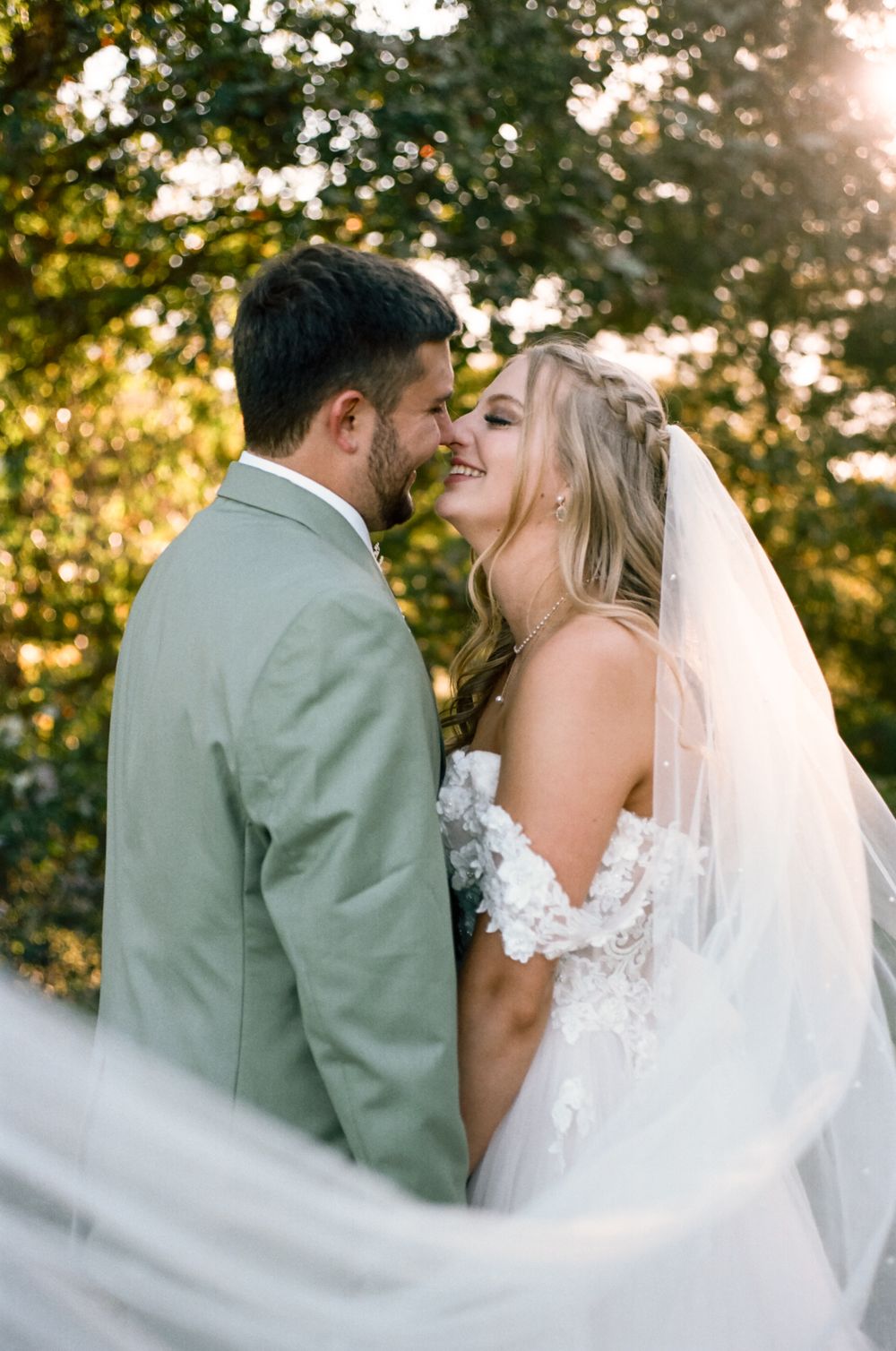 Intimate wedding moment captured with flowing veil against golden sunlit trees.