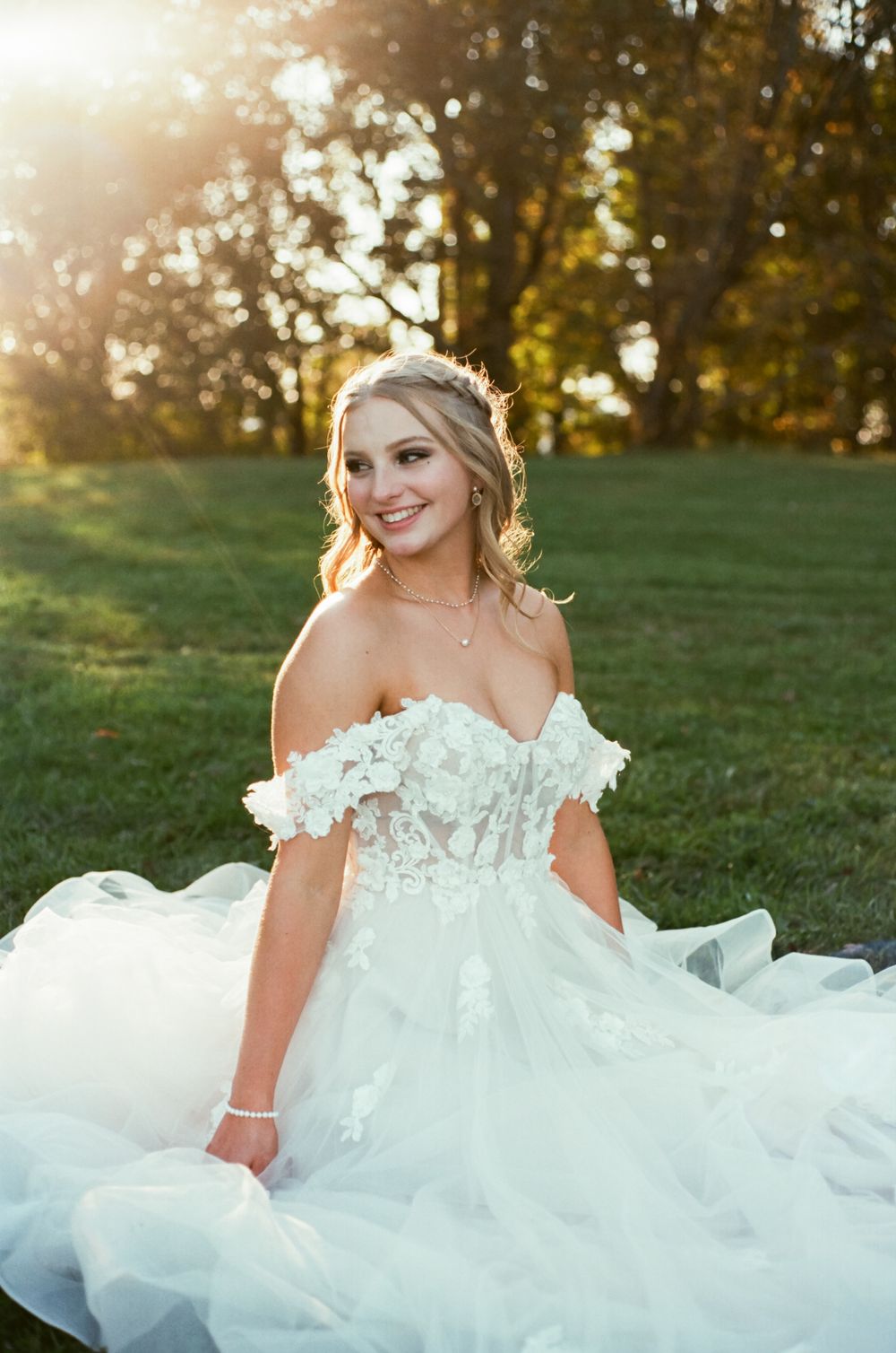 Bride in off-shoulder white gown poses outdoors during golden hour with a radiant smile.