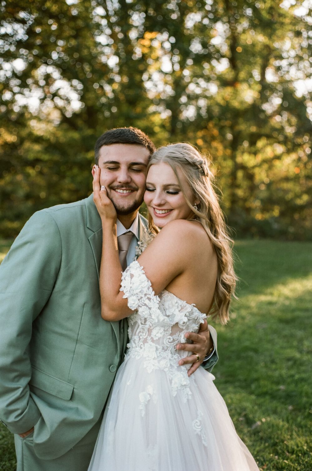 Joyful wedding couple sharing tender moment in sage green suit and lace wedding gown.