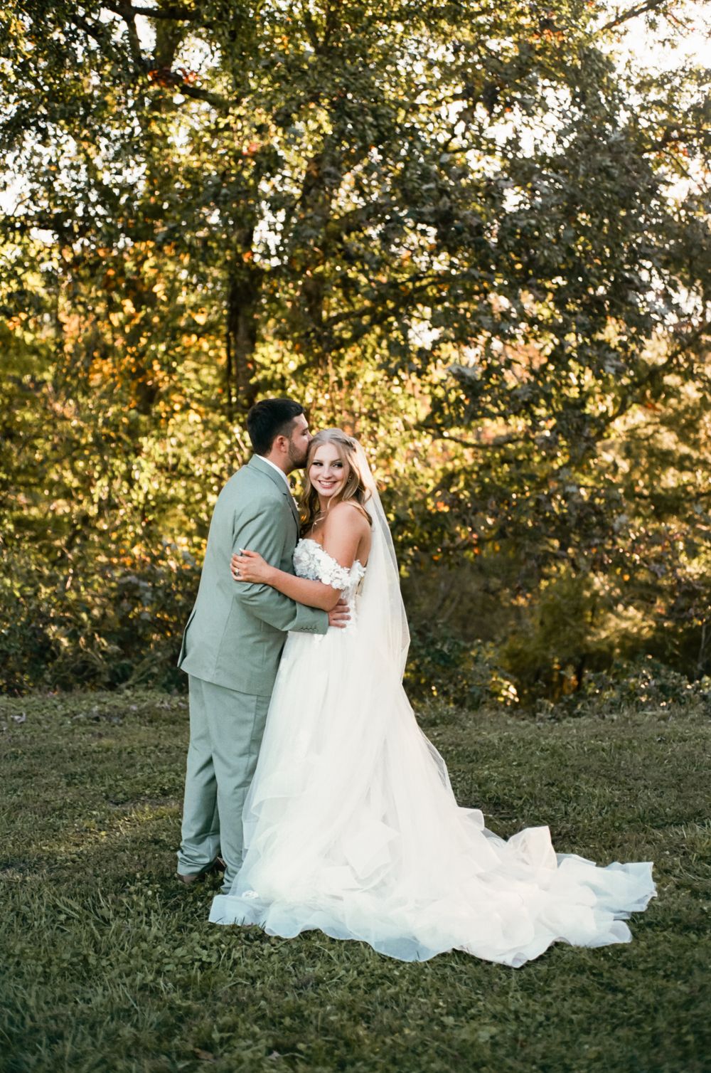 Elegant full-length wedding portrait with flowing white dress train against autumn trees.