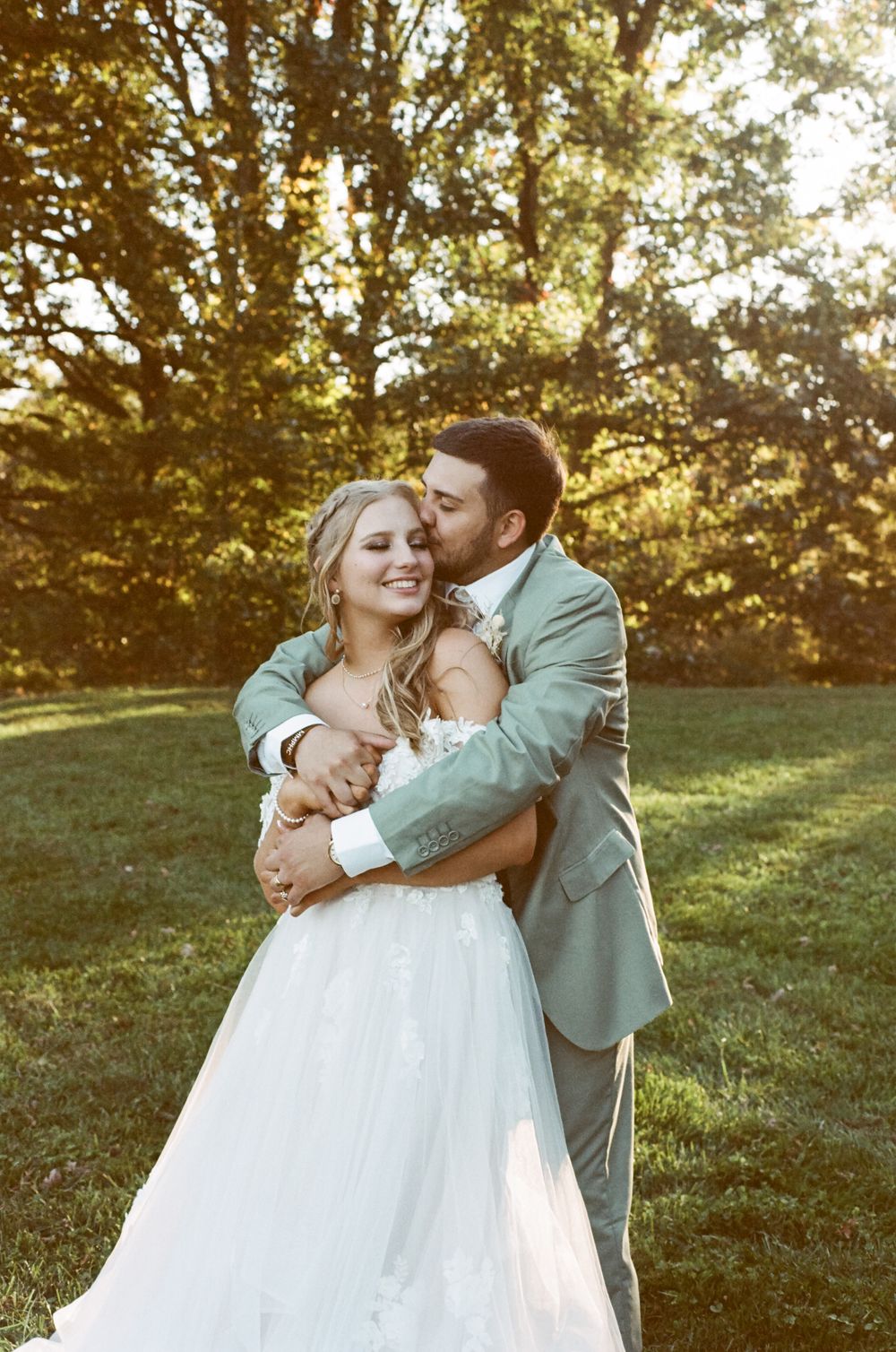 Natural outdoor wedding photo with couple embracing against sun-dappled foliage.