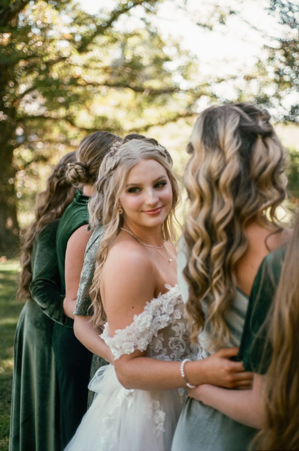 Bride in lace dress shares moment with bridesmaids wearing green dresses outdoors.